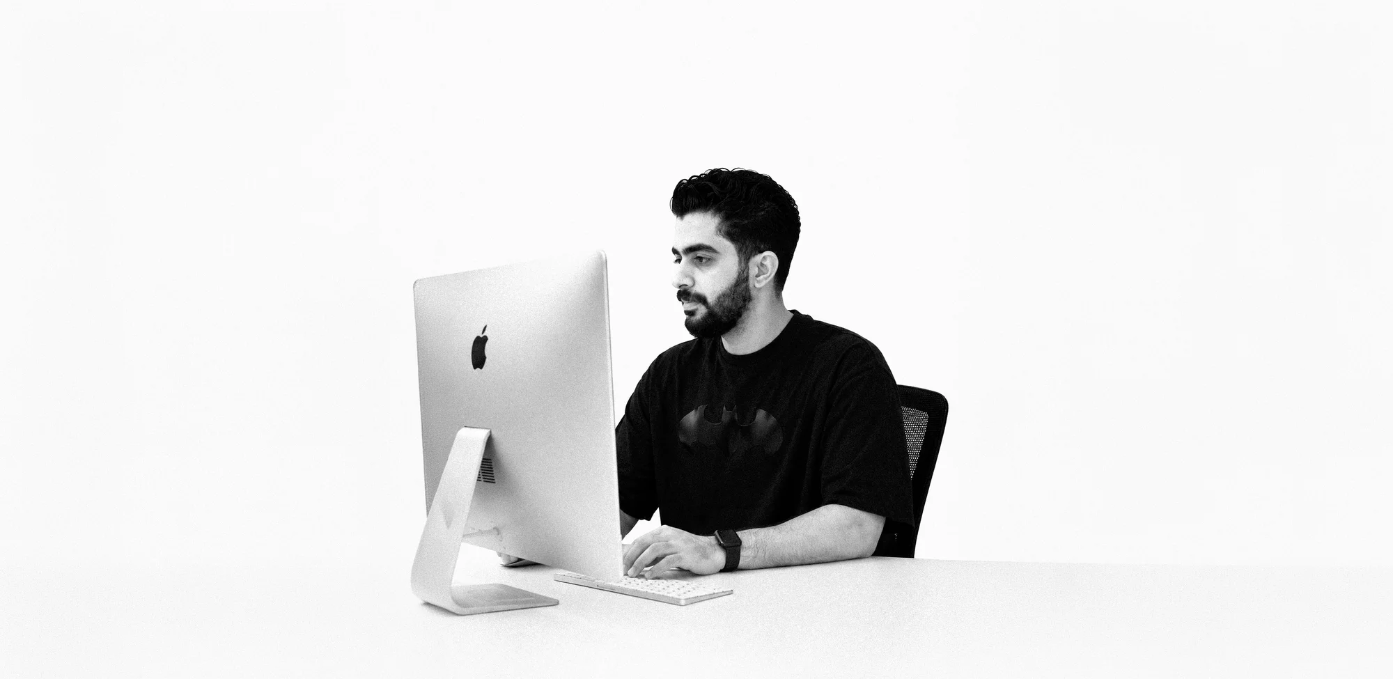Mohamed Gamal at work — seated at an iMac against a white studio ground, in black and white.