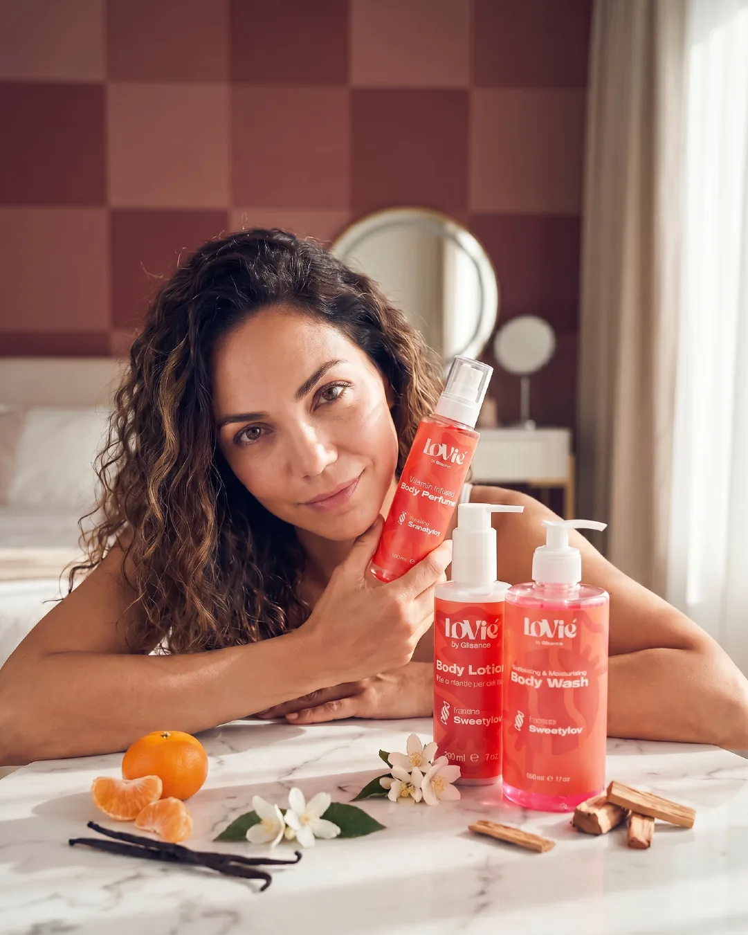 Portrait static: a woman lying on a bed holding the red Sweetylov perfume, the wash and lotion beside oranges, cinnamon and blossom.