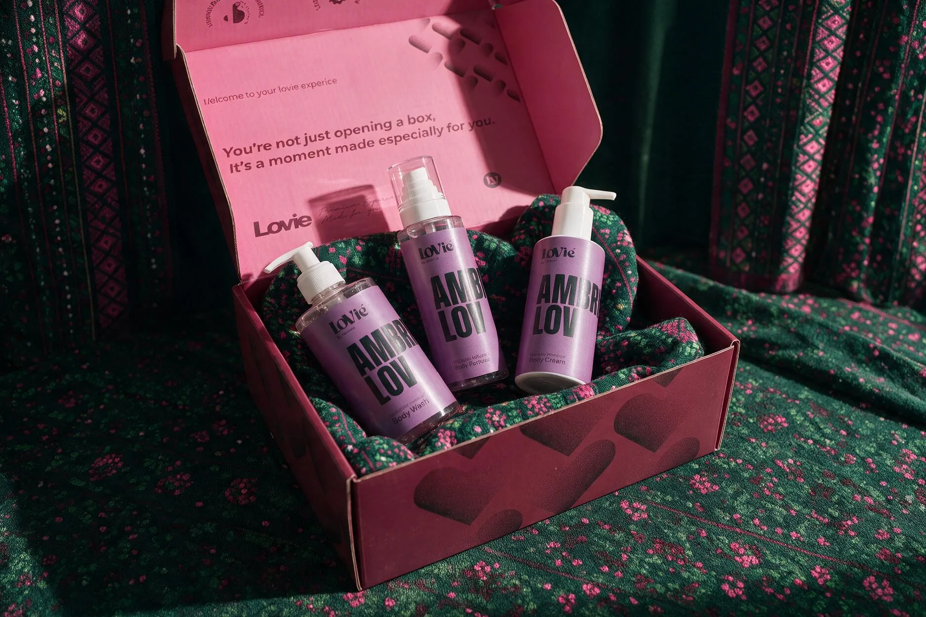 A pink LoVié box open on green sadu-print tissue, three violet AmbryLov bottles nested inside, a welcome line printed in the lid.