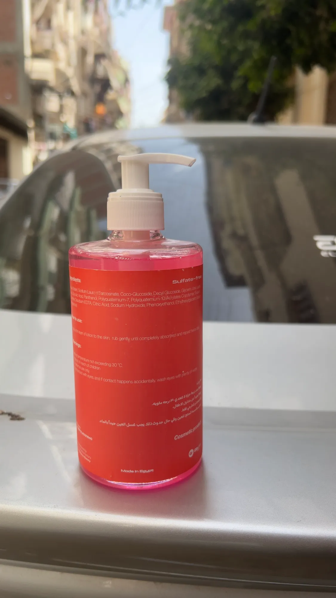 A printed red Lovie body wash bottle, Arabic back label out, standing on a white car roof in a Cairo street.