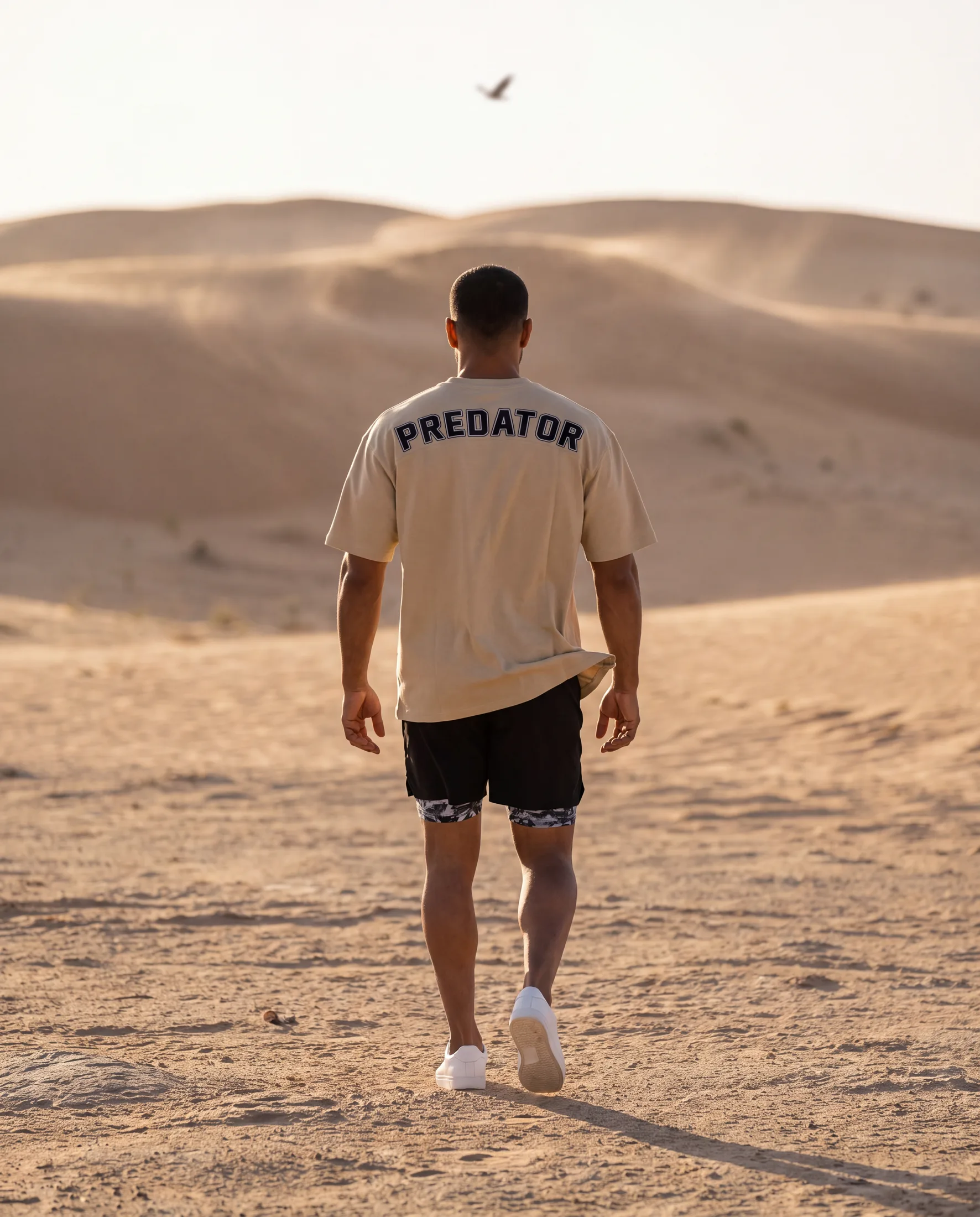 The athlete walking away from camera into the dunes, PREDATOR printed across the back of his sand-coloured tee.