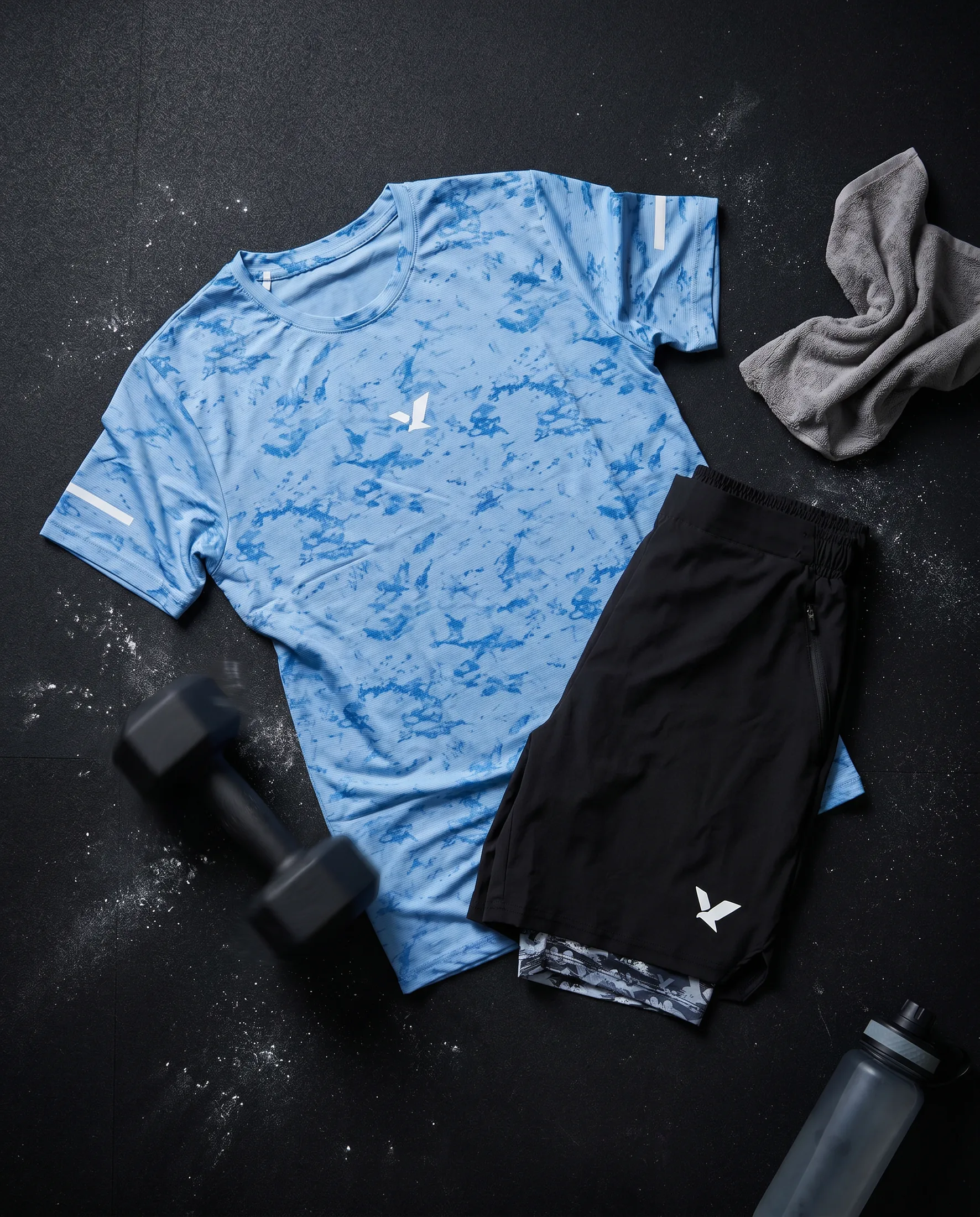 Flatlay of the pale blue OQAB tee and black shorts with camo liner, dumbbells and a towel on a dark ground.