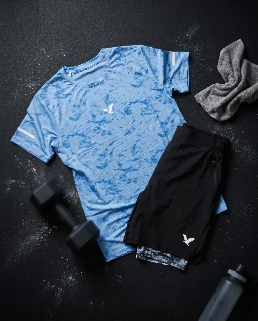 Flatlay of the pale blue OQAB tee and black shorts with camo liner, dumbbells and a towel on a dark ground.